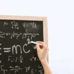 First Term Physics Examination for SSS 1 2 Close-up of hand writing physics equations on a chalkboard indoors.